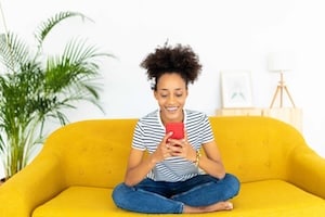 Happy mixed race young woman using mobile phone - Millennial african american girl teenager chatting on social media application while resting on sofa at home - Ethnicity and youth concept