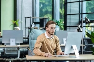 Serious and scorn-centered man in the office at work with a computer, man typing thoughtfully on keyboards, businessman young blond in sweater indoors loft, with green colors, programmer at work