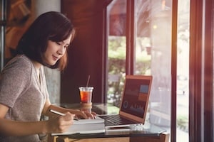 Isolated mockup image of laptop and Young business woman in casual dress sitting at table in cafe and writing in notebook.