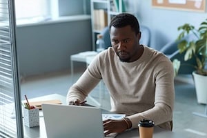 Focused black man using laptop in office behind glass partition wall