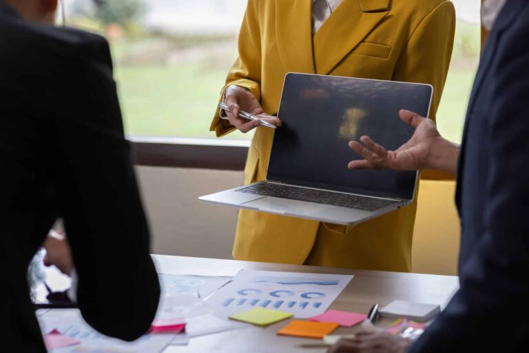 Marketer in yellow blazer presenting a blank laptop screen during website maintenance strategy meeting