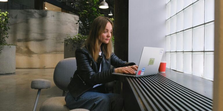 Woman working on laptop by a window in a modern workspace, representing a professional and reflective digital presence