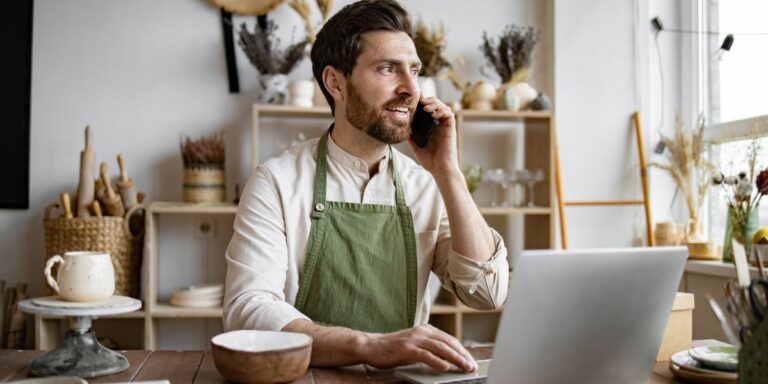 Mature Caucasian man in apron sits at table in ceramics shop and works with laptop. Experienced potter places online order while talking to customer over phone