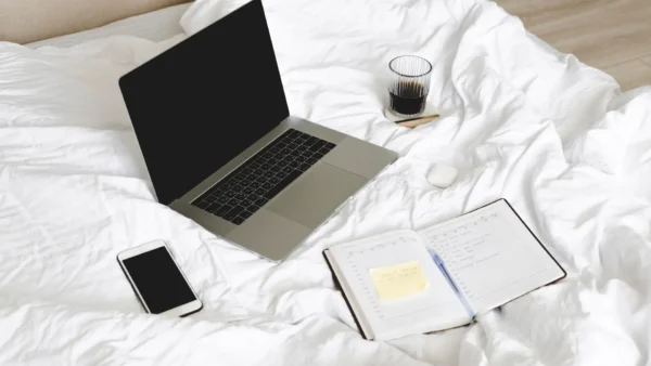 Open laptop and planner on a white bed, with coffee and phone nearby—an organized casual workspace
