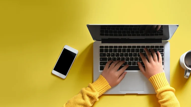 Hands typing on a laptop next to a phone and coffee cup on a bright yellow background