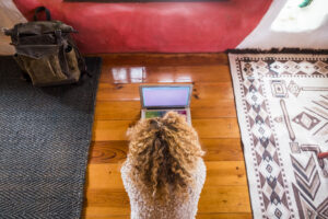 Overhead view of a woman with curly hair using a laptop on a wood floor, representing remote website content updates.