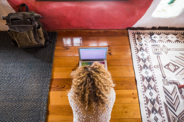 Overhead view of a woman with curly hair using a laptop on a wood floor, representing remote website content updates.