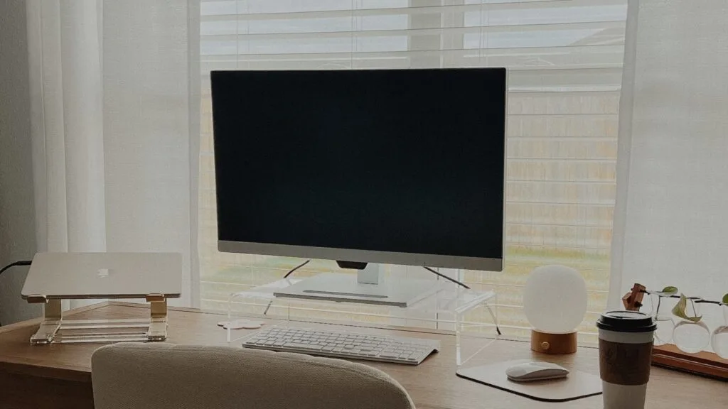 A clean home office setup with a desktop monitor, laptop, keyboard, lamp, coffee cup, and glasses on a wooden desk.