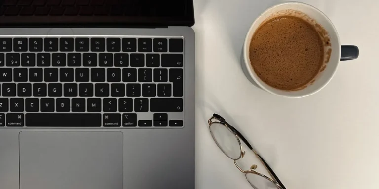 Laptop, coffee, and glasses on a desk representing the process of writing and managing consistent content when blogging for your website.