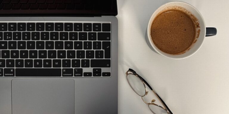Laptop, coffee, and glasses on a desk representing the process of writing and managing consistent content when blogging for your website.