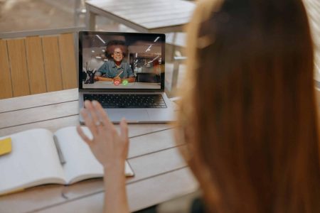 Back view of businesswoman talking with client via video call and waving Hi sitting in cafe terrace thought-leader
