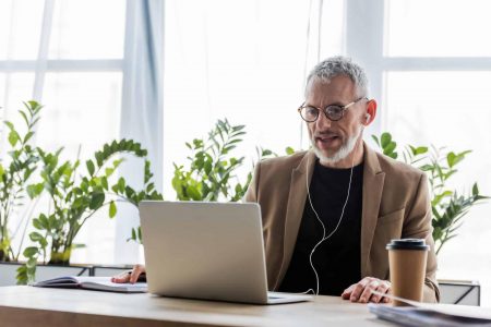 handsome businessman in glasses and earphones looking at laptop near paper cup on table marketing-budget
