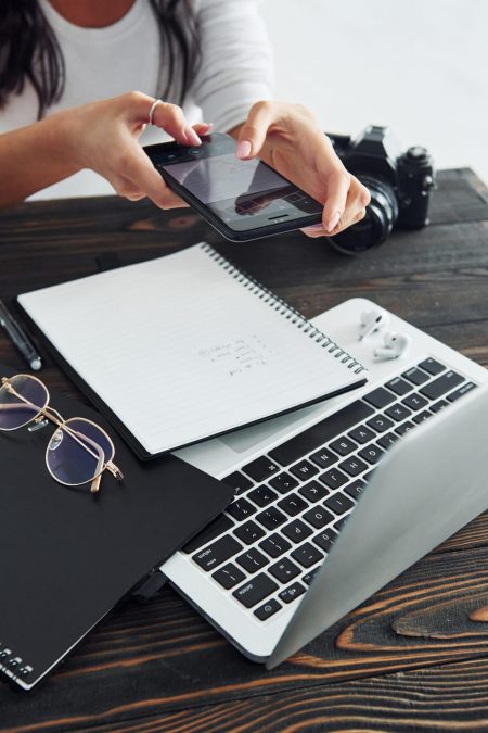 Holding phone. Young female freelancer working indoors in the office at daytime. Laptop and notepad personalize